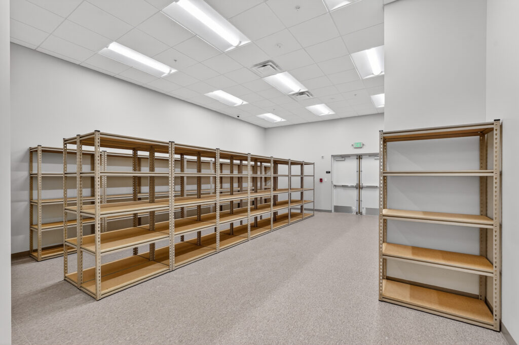 Empty metal shelving units arranged in an L-shape inside a room with white walls, a drop ceiling, and double exit doors.
