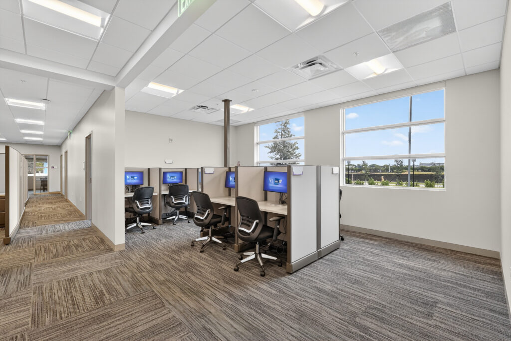 Office space with cubicle desks featuring computers and rolling chairs, next to a windowed wall.