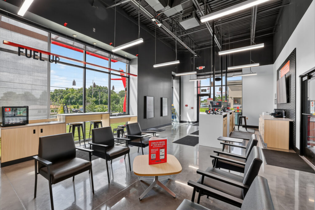 Interior of a modern auto shop waiting area with black leather chairs, a light wood coffee table, a service counter, and floor-to-ceiling windows.