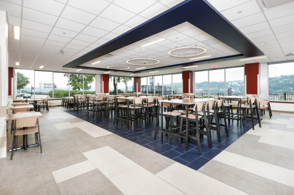 A large cafeteria with rows of high-top wooden tables and chairs on a blue and white tiled floor, surrounded by floor-to-ceiling windows.