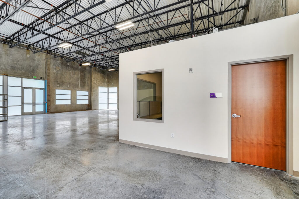 An open warehouse space with concrete floors, a glass-paneled entry, and an enclosed office room with a wood door and a window.