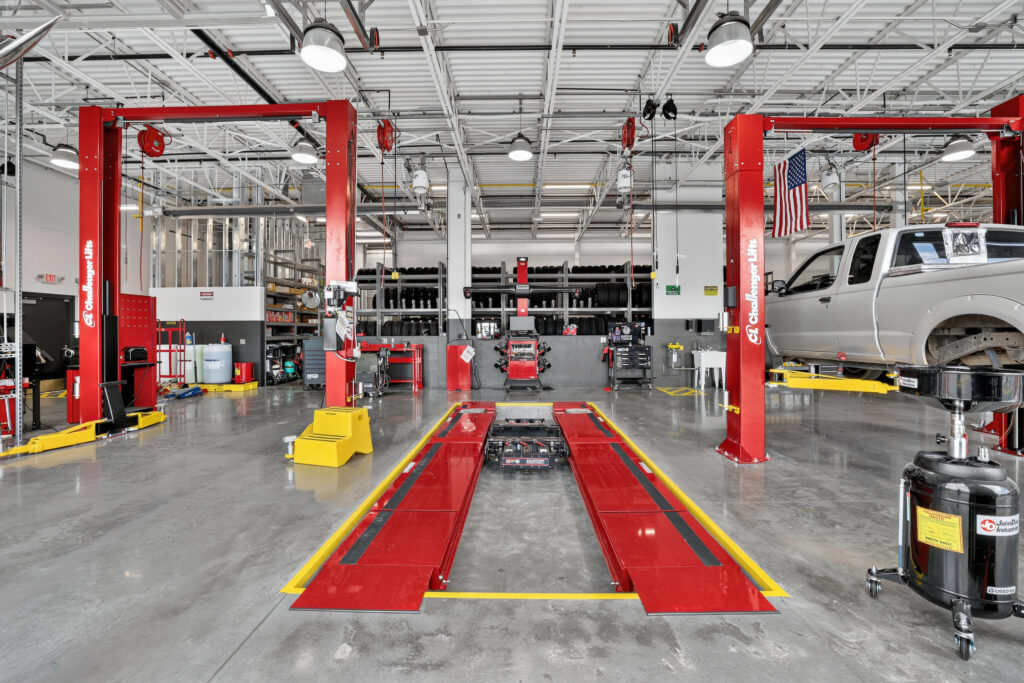 An automotive repair shop featuring two red vehicle lifts, one holding a silver pickup truck, with racks of tires in the background.