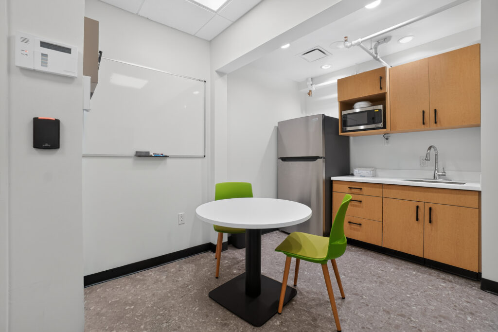 Small office breakroom with a round table, two green chairs, a stainless steel refrigerator, and wooden cabinets above a sink.