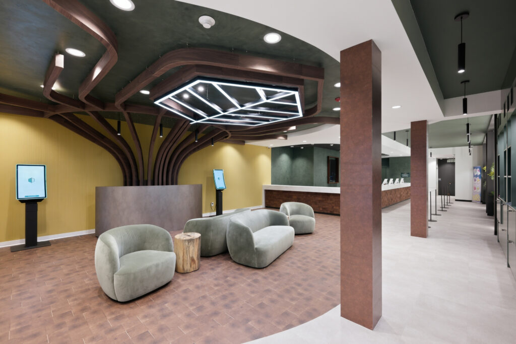 Lobby with curved grey sofas, a wooden tree-like ceiling feature with geometric lighting, two digital kiosks, and a reception desk in the background.