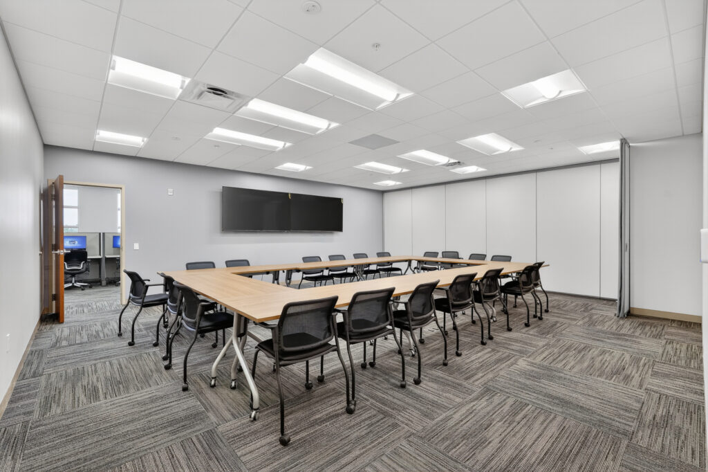 A U-shaped conference table surrounded by black mesh chairs in a grey-walled meeting room with a wall-mounted monitor.