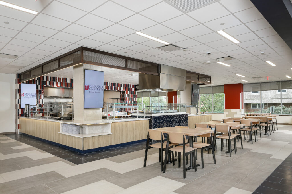 A modern dining hall serving area with food stations, a digital menu board for Duquesne University Dining Services, and several wooden dining tables with chairs.