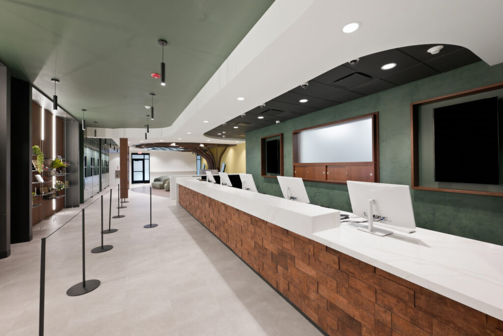 A long reception desk with white countertops and a textured brick base, featuring several computer monitors and a queueing area with floor stanchions.
