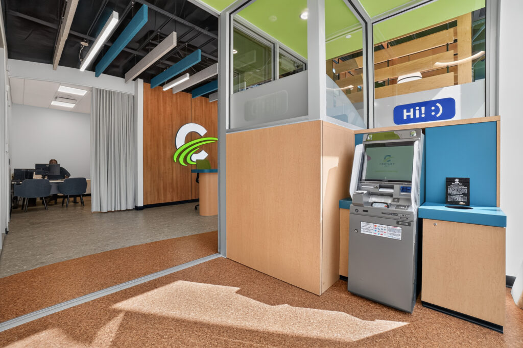 An ATM kiosk inside a modern office lobby with a wood-paneled wall, a reception desk, and a person working at a computer in the background.