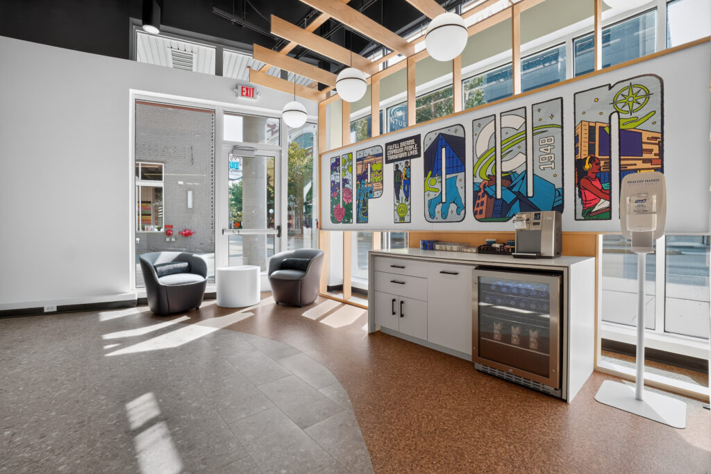 A waiting area with two lounge chairs, a beverage station with a glass-front refrigerator, and a decorative wall panel featuring colorful illustrations and text.