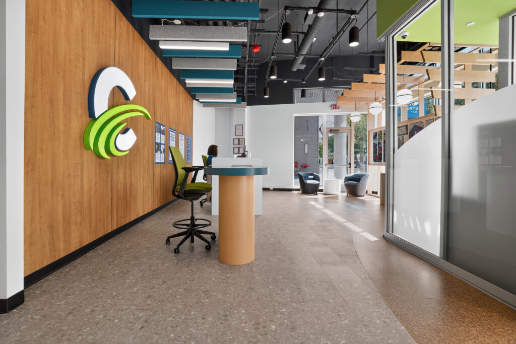 Modern office reception area with a wooden wall featuring a large green and white “C” logo, a service desk, and a person sitting at a computer.