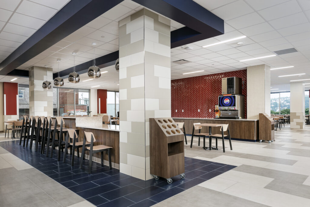 Modern cafeteria interior with a long counter, bar seating, tables, and a Pepsi soda fountain against a red tiled wall.