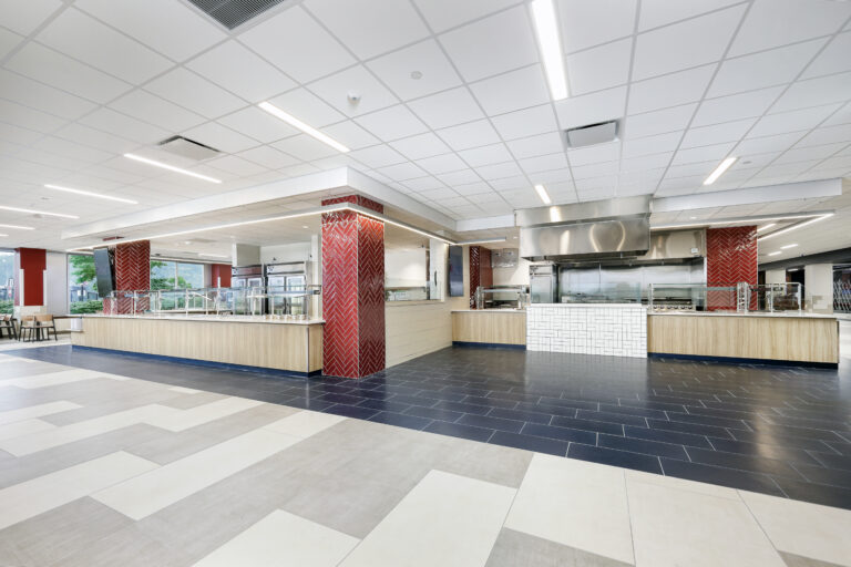 An empty cafeteria serving area with long counters, stainless steel equipment, and red herringbone tile columns.