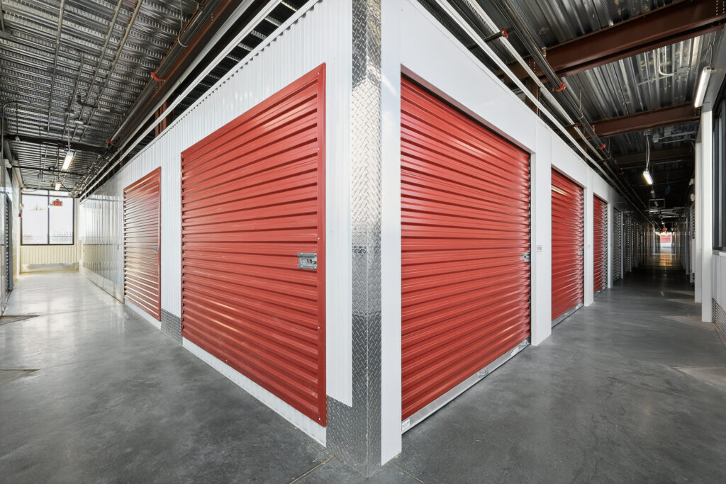 A corner of a storage facility featuring rows of red roll-up metal doors in a concrete hallway.