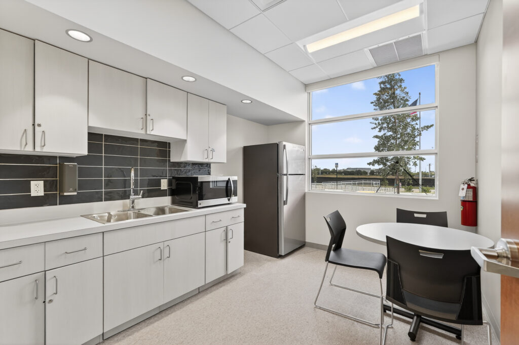 Office breakroom with white cabinets, a sink, microwave, refrigerator, and a small round table with two chairs near a window.