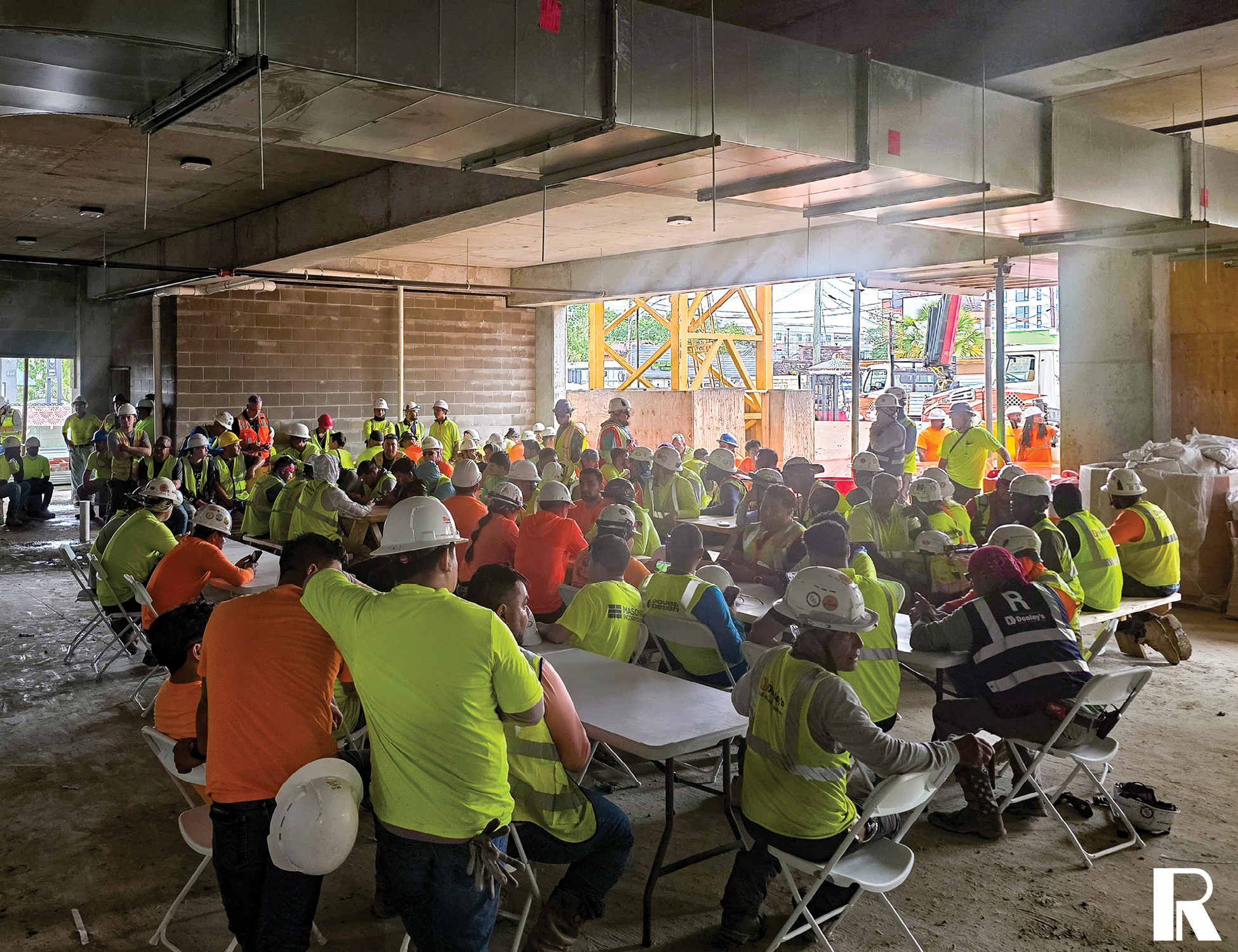 Large group of construction workers in hard hats and high-visibility vests seated at tables for a meeting in an unfinished building.