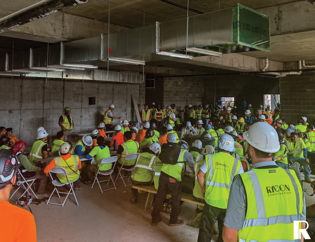 A large group of construction workers in hard hats and high-visibility vests attending an indoor meeting at a job site.