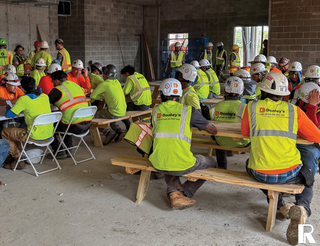 Construction workers in hard hats and high-visibility vests sitting at tables and benches in an unfinished building.
