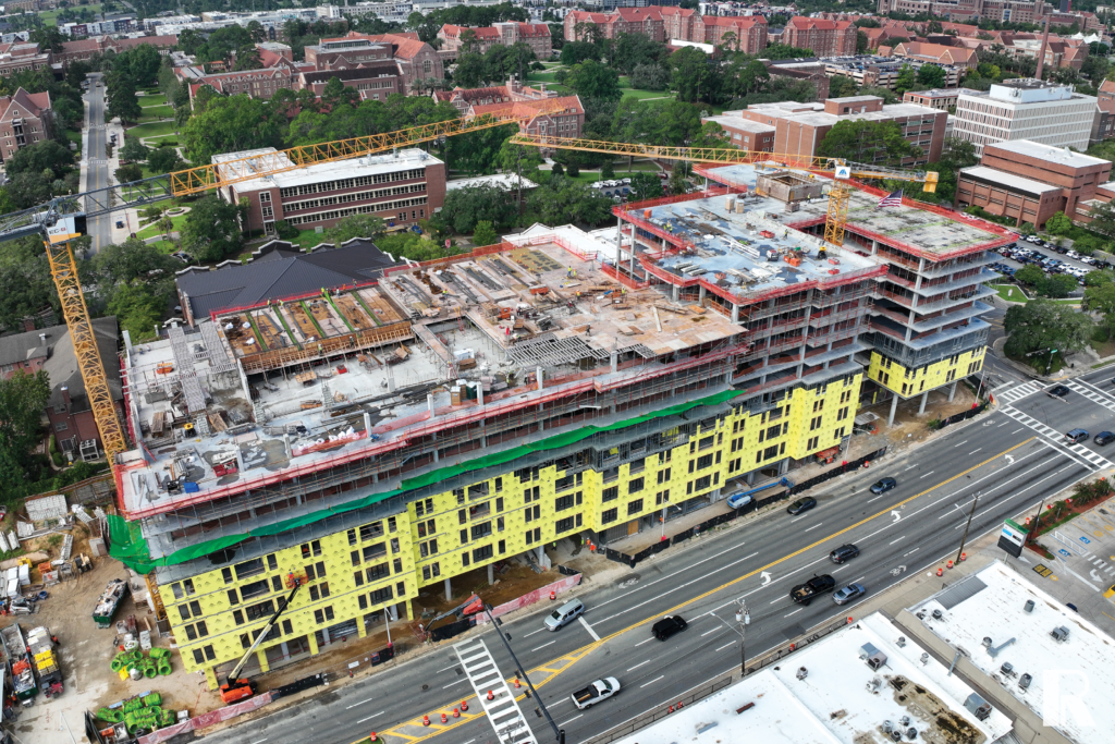 Aerial view of a large multi-story building under construction with yellow exterior panels and a yellow tower crane, situated next to a busy multi-lane road.