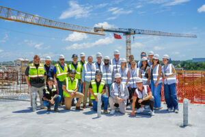 A large group of construction workers and project team members wearing hard hats and high-visibility vests posing for a photo on a rooftop construction site with a crane in the background.
