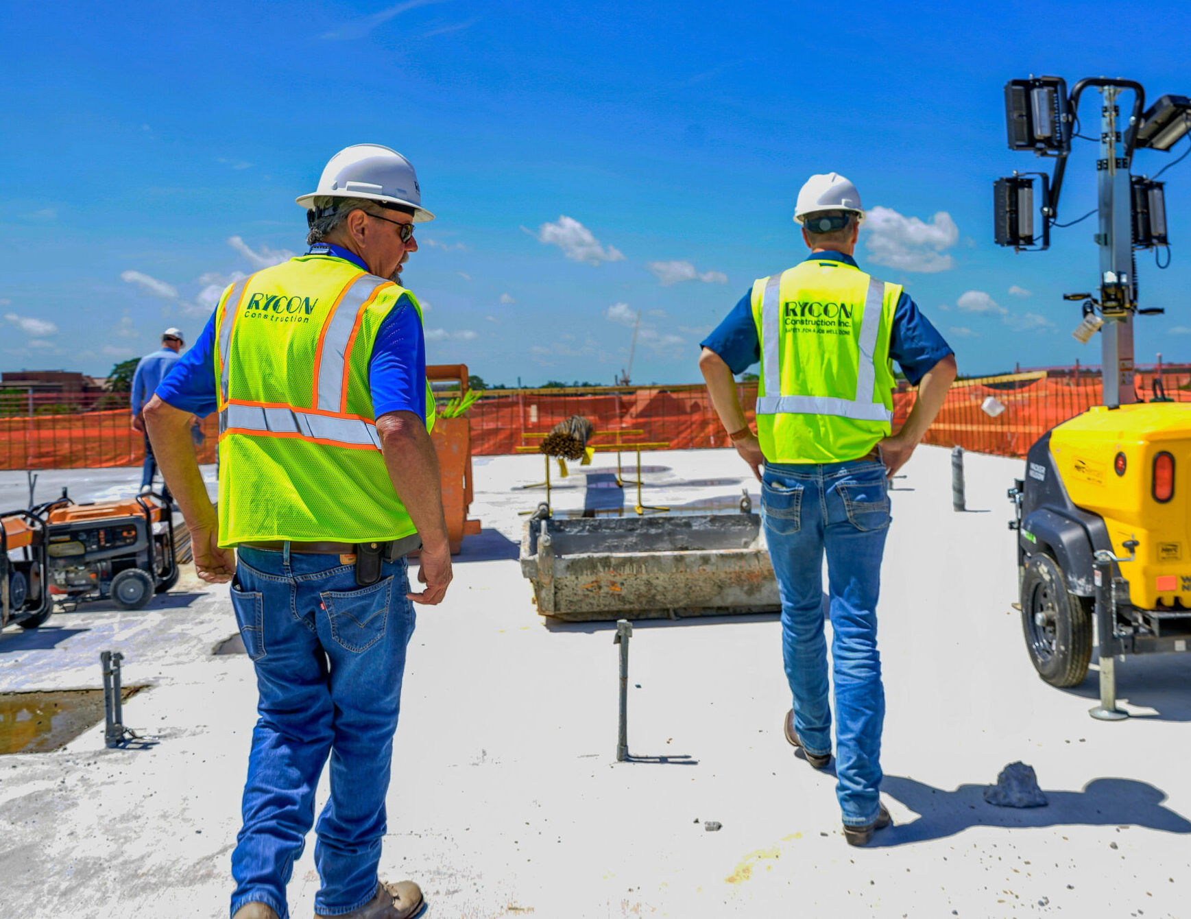 Two construction workers wearing hard hats and high-visibility vests walk across a flat roof at a job site.