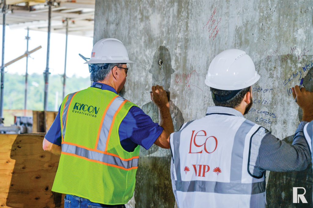 Two construction workers in hard hats and high-visibility vests writing their names on a concrete wall.