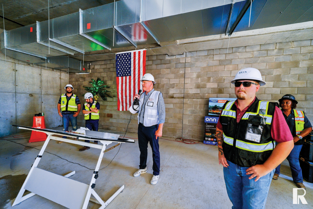 A man in a white hard hat speaks into a microphone at an indoor construction site, with three other workers standing nearby and an American flag hanging on the block wall behind him.