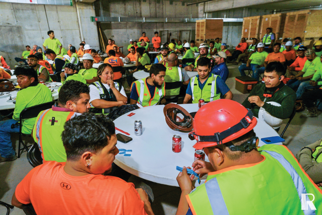 Construction workers in high-visibility vests sit at circular tables in a large concrete room, eating and socializing during a break.