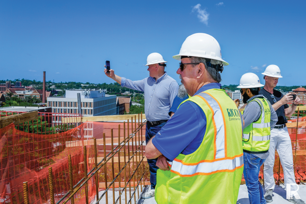 Four construction workers in hard hats stand on a building rooftop, with one person holding up a phone to take a photo of the cityscape.