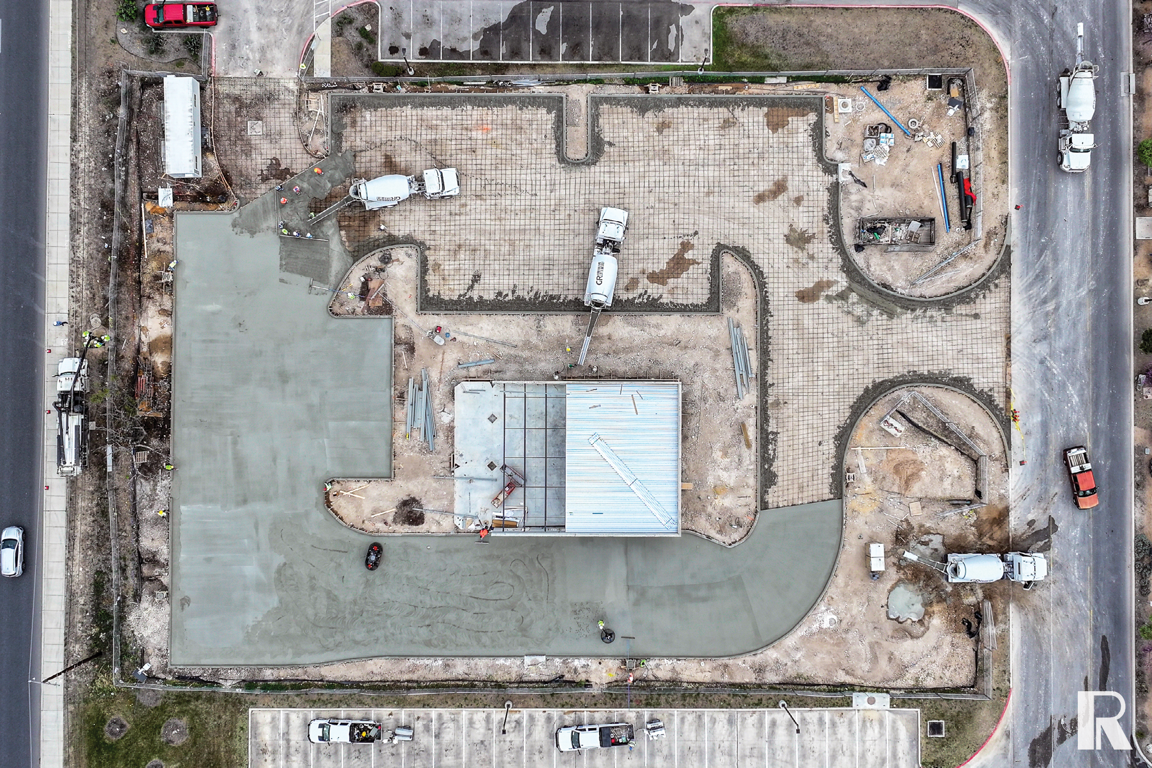 Aerial view of a construction site showing workers pouring concrete into a prepared rebar foundation grid, with multiple cement mixer trucks positioned around the site.