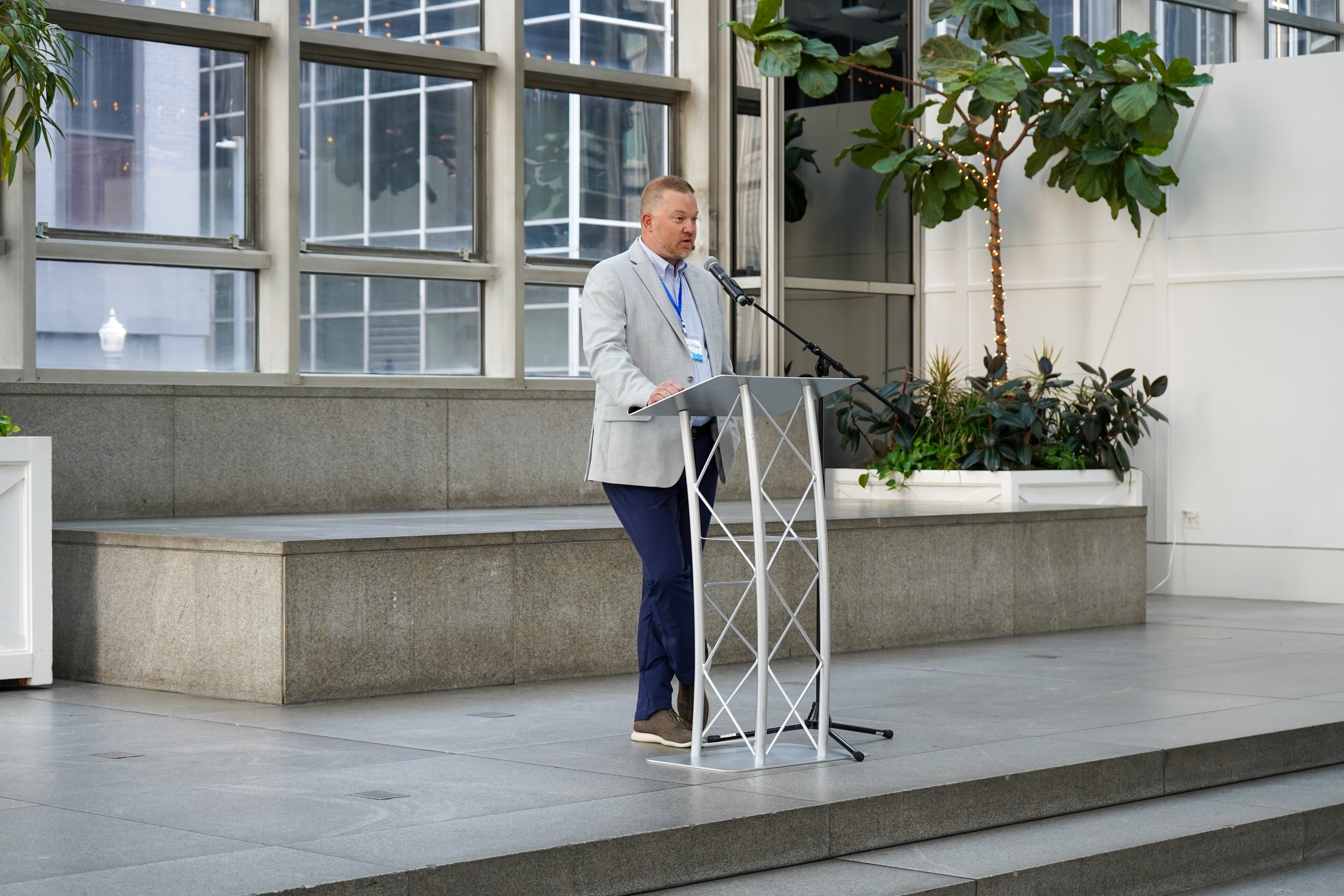 A man in a light gray blazer speaks at a podium in an office atrium.