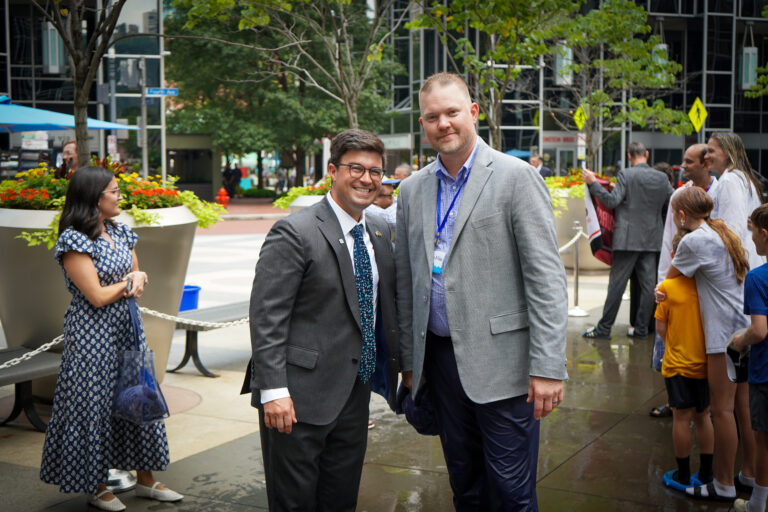 Two men in business attire stand side-by-side outdoors, with other people and an urban plaza visible in the background.