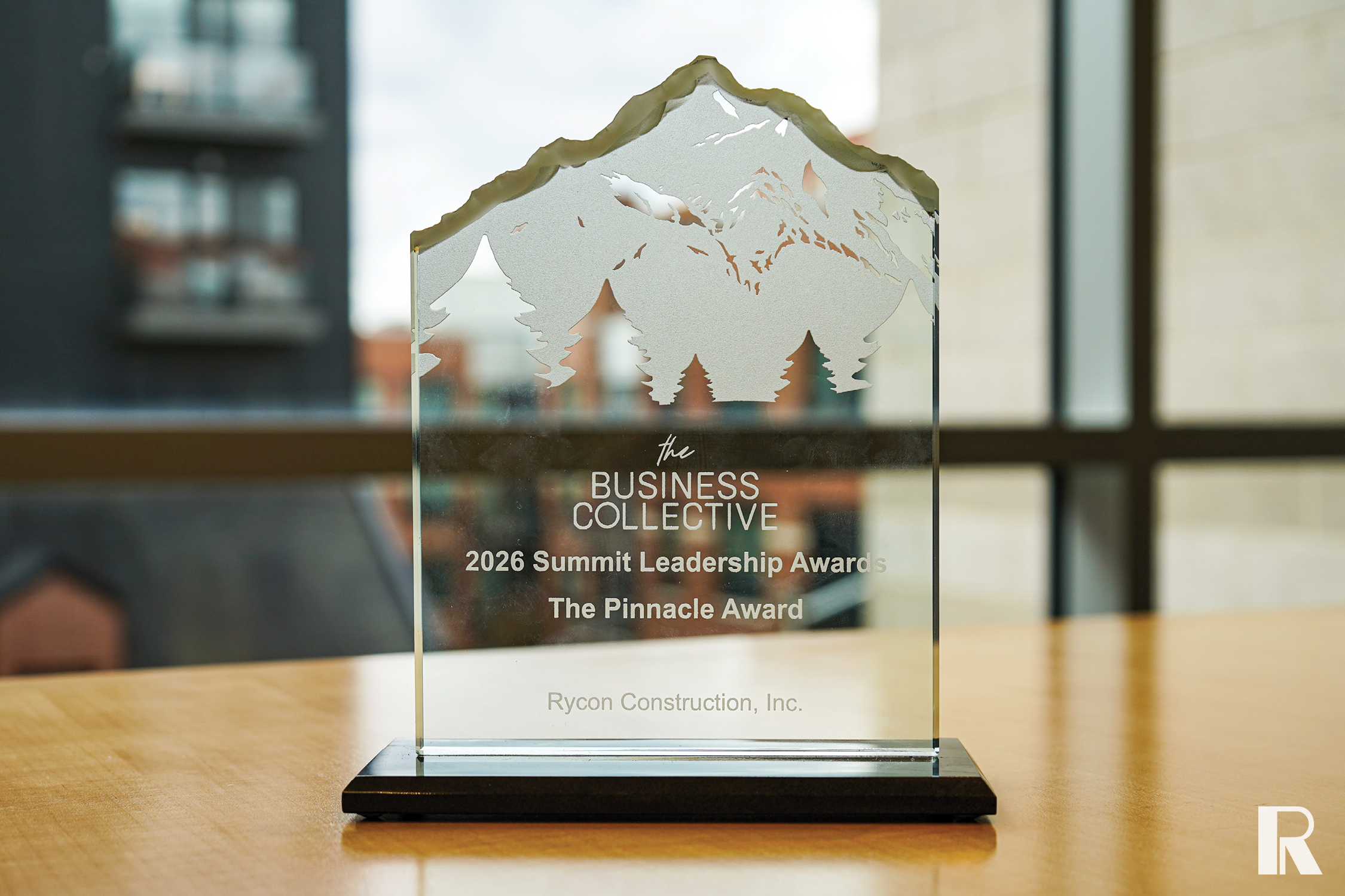 Glass mountain-shaped trophy engraved with “The Business Collective 2026 Summit Leadership Awards The Pinnacle Award” and “Rycon Construction, Inc.” on a wooden table.