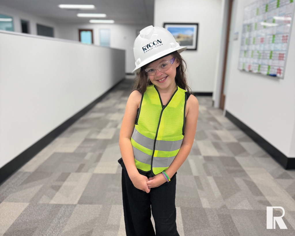 A young girl in an office hallway wearing a Rycon Construction hard hat, safety glasses, and a neon yellow safety vest.