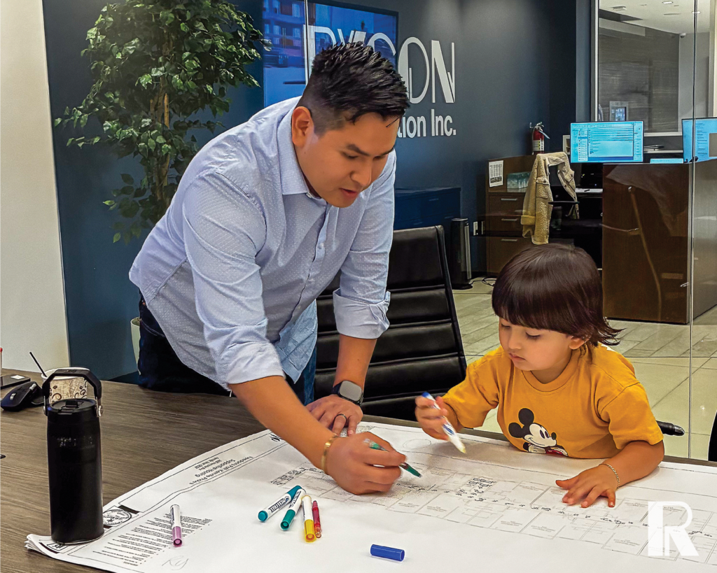 A man in a light blue shirt and a child in a yellow shirt draw on a large architectural blueprint spread across a desk.
