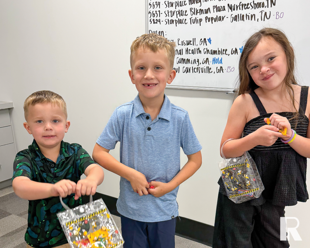 Three children holding small, clear plastic gift bags in front of a whiteboard with handwritten location names.