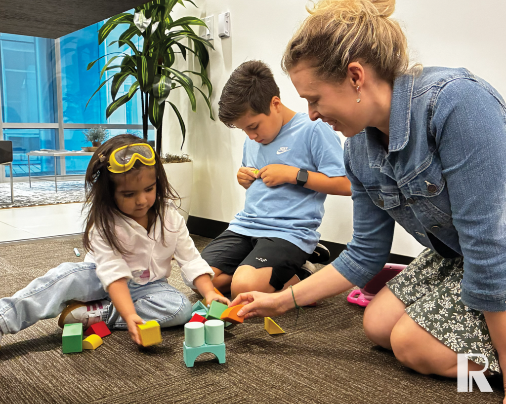 A woman and two children sit on a floor playing with colorful wooden building blocks.