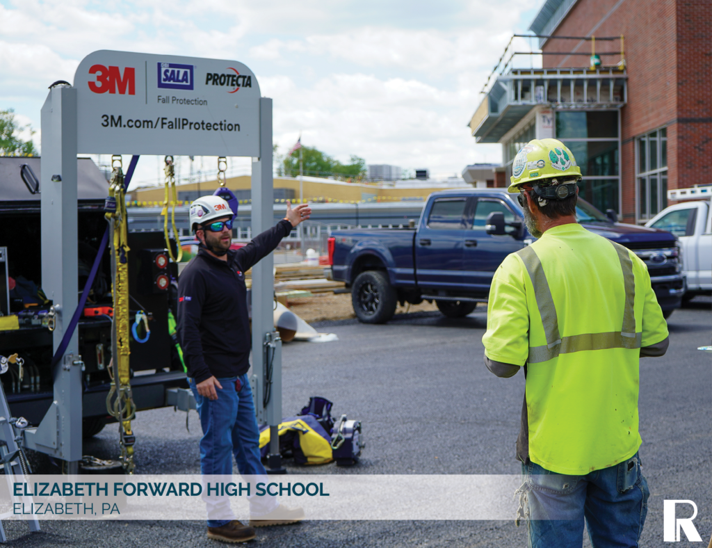 Two construction workers stand near a 3M mobile fall protection unit in a parking lot.