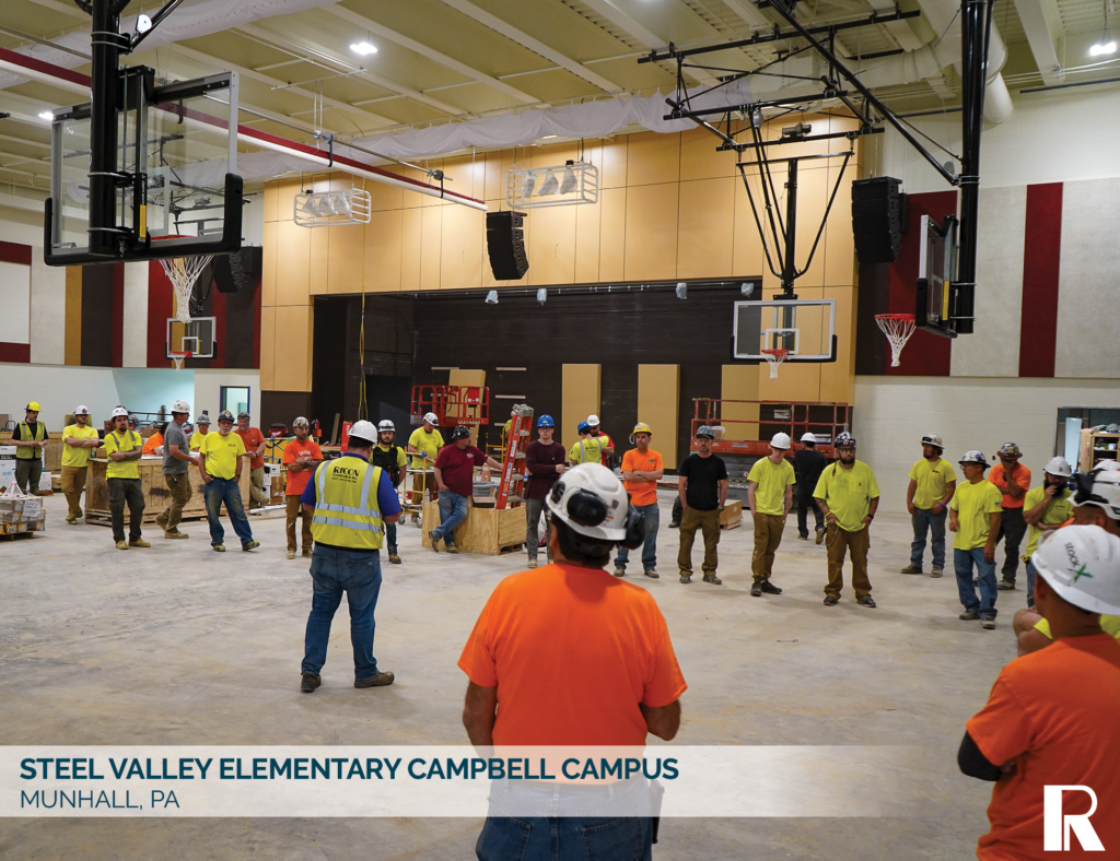 A group of construction workers in hard hats and high-visibility gear stand on the floor of a school gymnasium during a meeting.