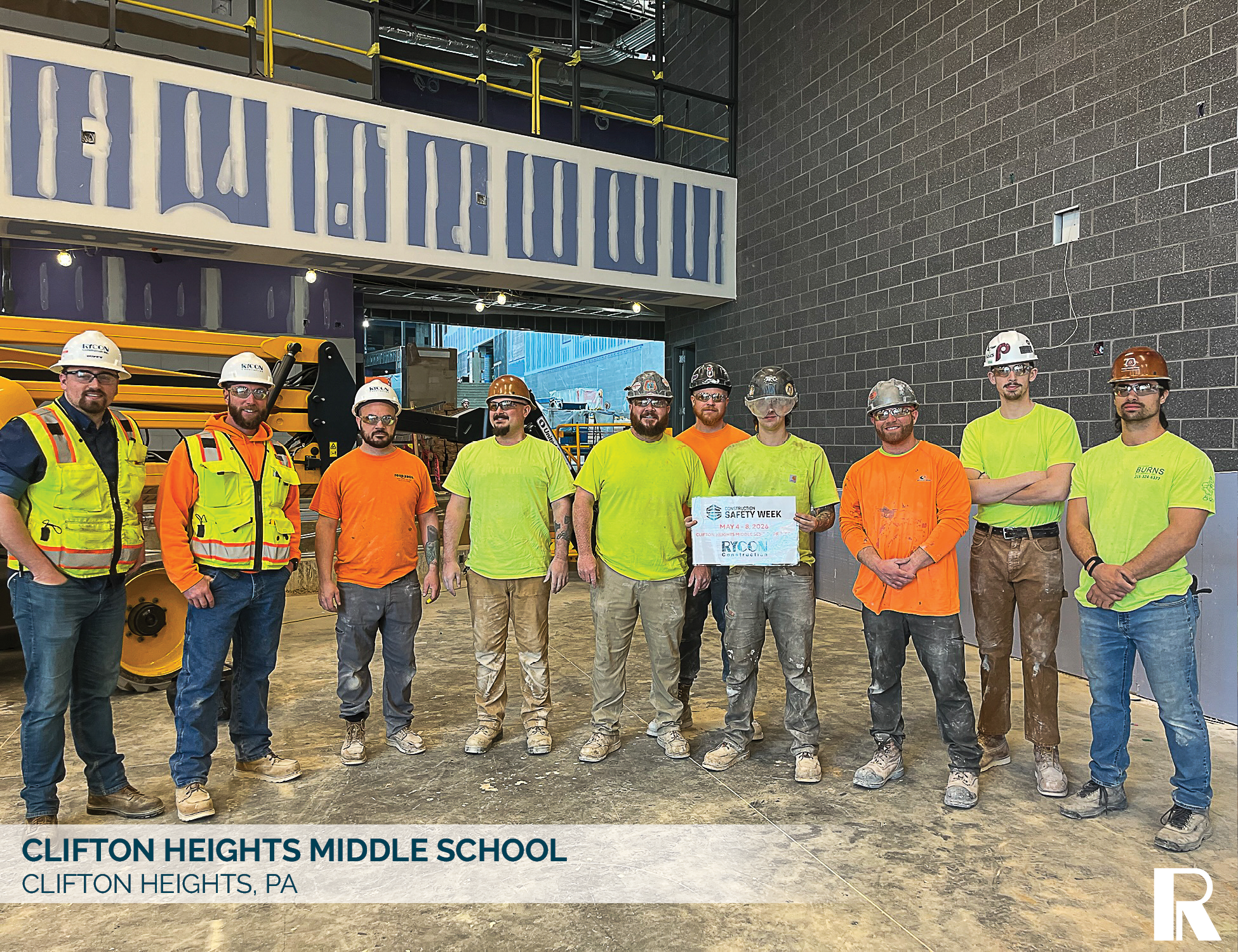 A group of eleven construction workers in hard hats and high-visibility clothing standing on a job site at Clifton Heights Middle School, with one worker holding a Construction Safety Week sign.