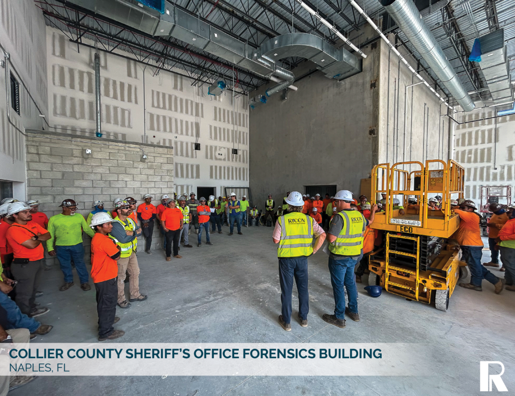A group of construction workers in hard hats and safety vests stand in a large, unfinished building with exposed industrial ceiling ducts and a yellow scissor lift.