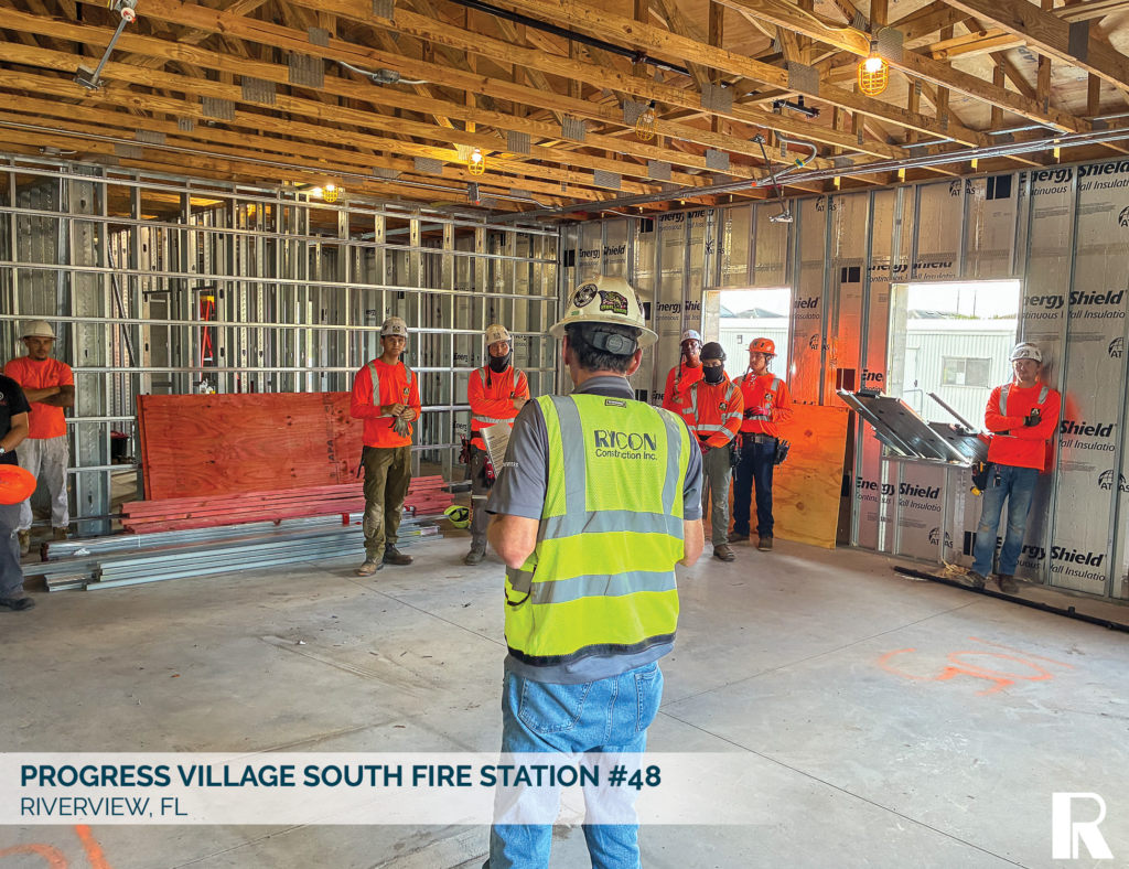 A construction supervisor in a high-visibility vest addresses a crew of workers wearing hard hats and orange shirts inside an unfinished building framed with metal studs.