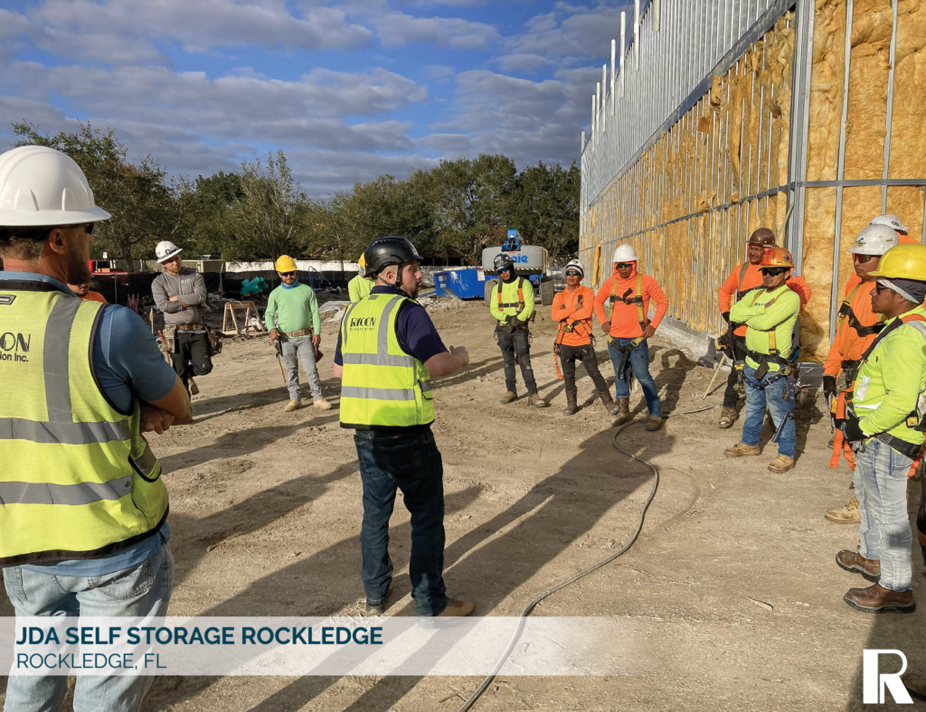A project manager in a high-visibility vest speaks to a group of construction workers at a job site with a half-built building in the background.