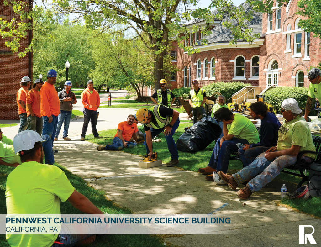 A group of construction workers in high-visibility vests and hard hats gathered on a university campus lawn.