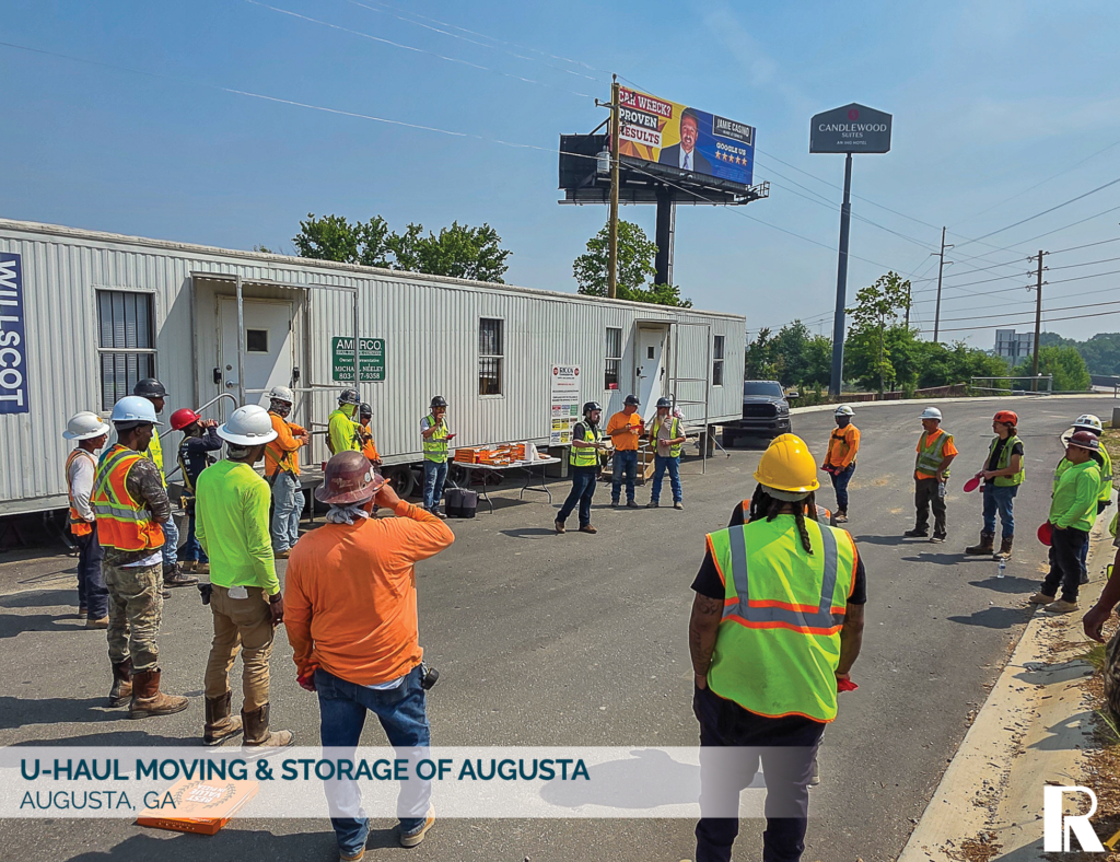 A group of construction workers in high-visibility vests and hard hats standing in a circle for a morning meeting in front of a portable office trailer.