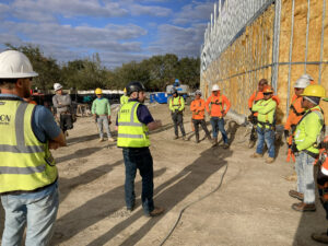 A group of construction workers in hard hats and high-visibility gear gathered for a meeting on a job site next to a building under construction.