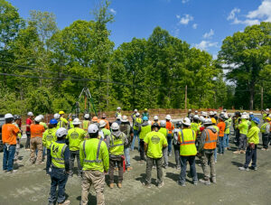 A large group of construction workers in hard hats and high-visibility vests gather at a job site near a wooded area.