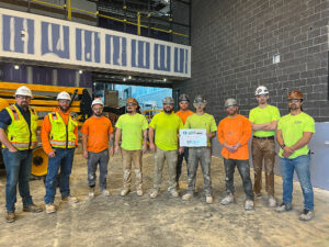 Ten construction workers wearing high-visibility gear and hard hats pose in an unfinished building, with one worker holding a Construction Safety Week sign.