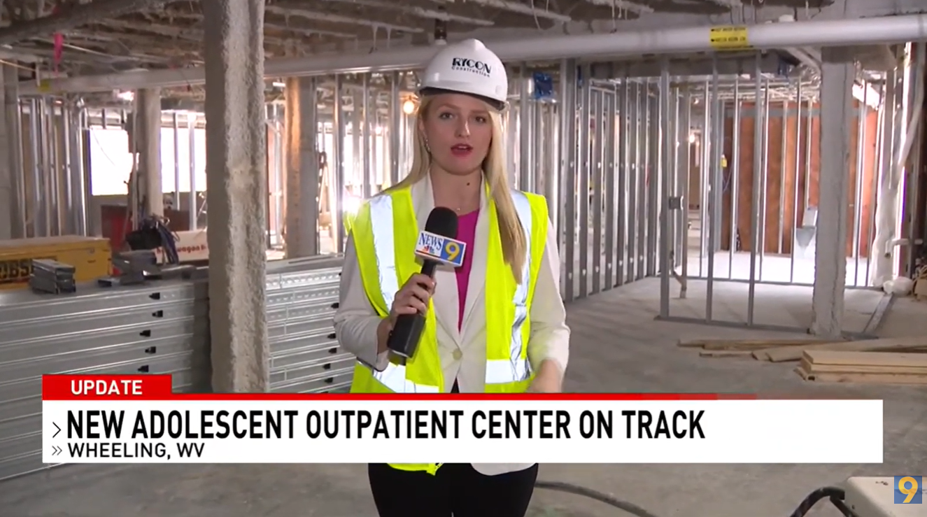 A news reporter in a hard hat and safety vest stands in a building under construction, with a chyron reading “New Adolescent Outpatient Center on Track.”