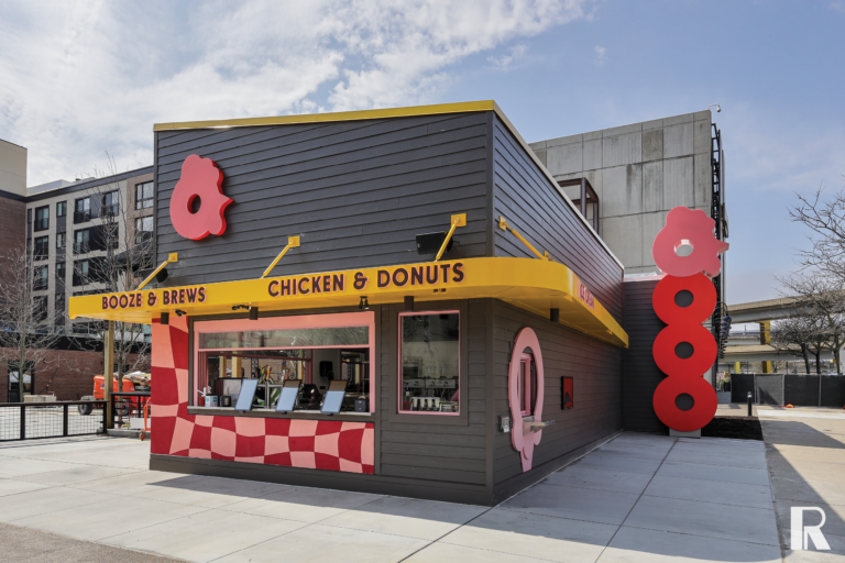 A small, dark gray walk-up restaurant with a yellow awning, red chicken-shaped signage, and text reading “Booze & Brews” and “Chicken & Donuts.”