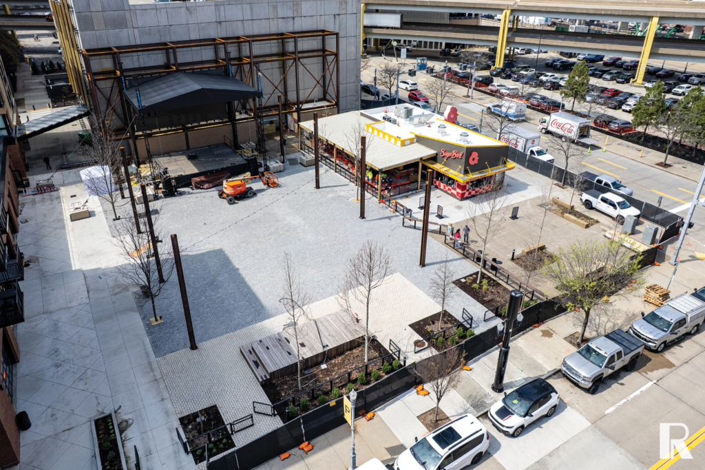 Aerial view of an outdoor plaza featuring a stage with scaffolding and a nearby Sugar Bowl restaurant, surrounded by parking lots and city streets.
